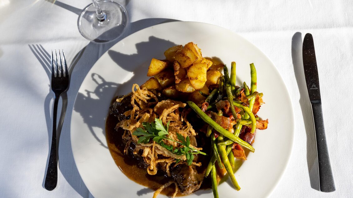 Ein Teller mit saftigem Fleisch, umgeben von grünen Bohnen, knusprigen Kartoffeln und einer leckeren Sauce. Das Gericht ist hübsch angerichtet und wird von einem Glas Wein begleitet. | © Lipizzanerheimat-Schiffer