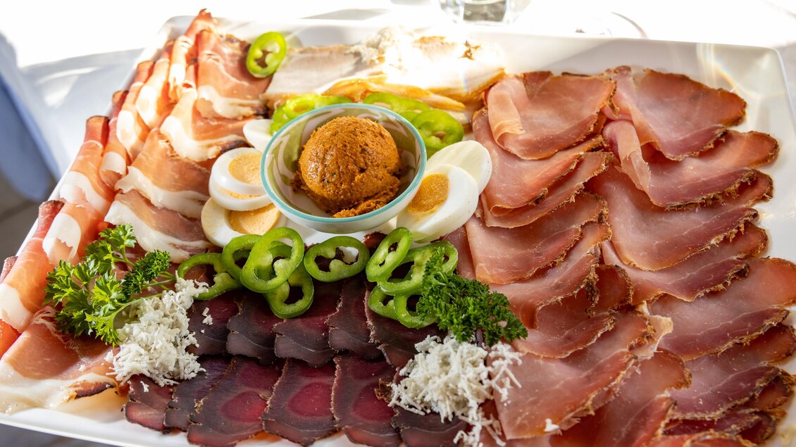 Eine Auswahl an verschiedenen Schinkensorten auf einem großen Teller. In der Mitte befindet sich eine Schüssel mit einer würzigen Paste und frischem Gemüse. | © Lipizzanerheimat-Schiffer