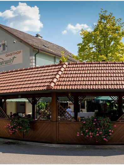 Ein gemütliches Restaurant mit einer überdachten Terrasse und bunten Blumen. Der Himmel ist klar und das Gebäude ist im traditionellen Stil gehalten. | © TV Region Graz - Lunghammer