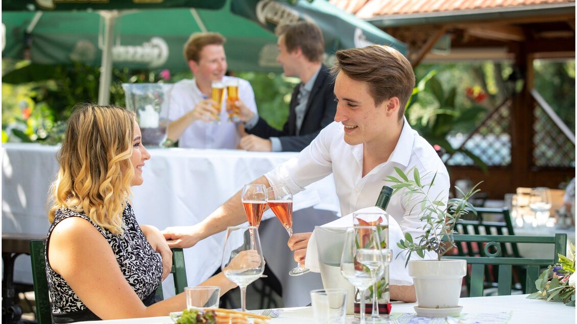 Ein elegantes Restaurant im Freien mit einer jungen Frau und einem jungen Mann, die auf einer festlich dekorierten Tafel anstoßen. Im Hintergrund sind weitere Gäste, die sich unterhalten und Getränke genießen. | © TV Region Graz - Lunghammer