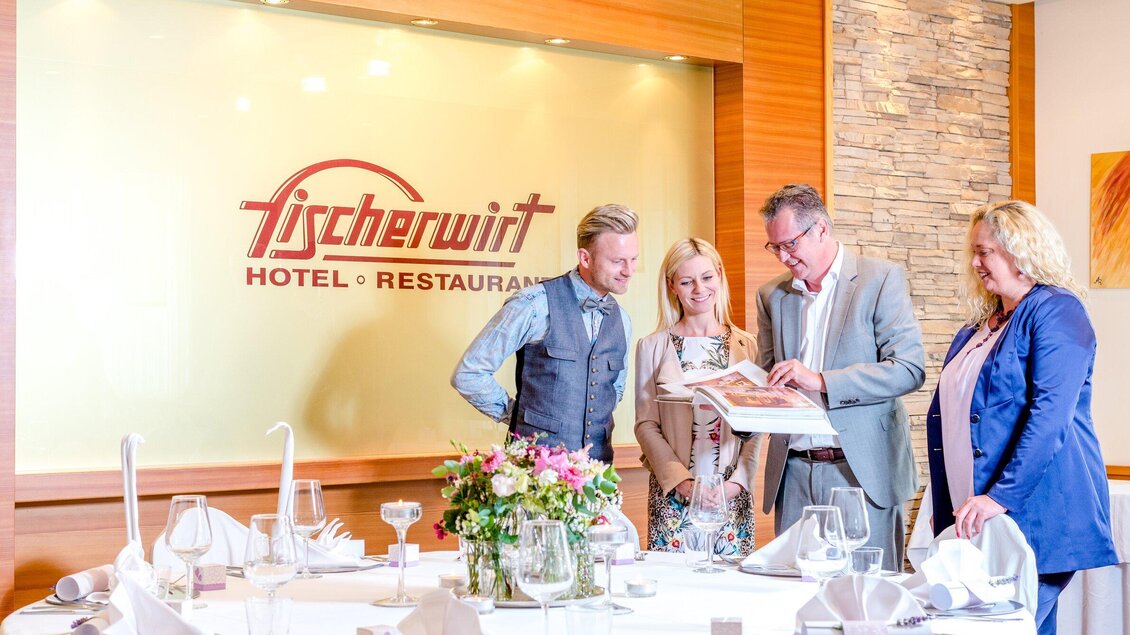 Eine Gruppe von Menschen steht an einem Tisch in einem stilvollen Restaurant. Sie betrachten eine Menükarte vor dem Logo des Hotels und Restaurants „Fischerwirt“. | © TV Region Graz - Mias Photoart