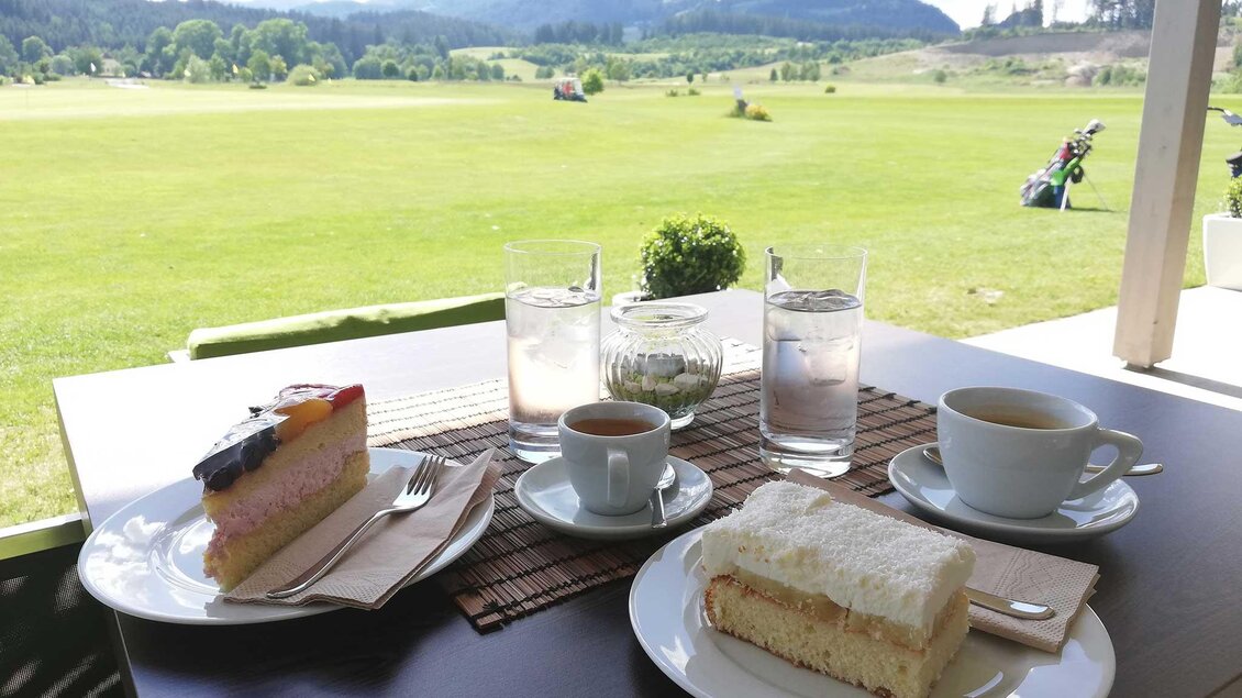 Ein Tisch mit zwei verschiedenen Kuchenstücken, Kaffee und Wasser. Im Hintergrund ist eine grüne Landschaft mit Hügeln und einem Golfplatz zu sehen. | © Restaurant - Bistro Golfblick