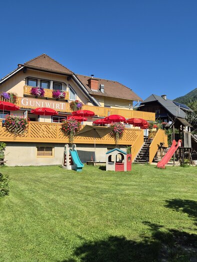 Ein hübsches Gebäude mit bunten Blumen und Sonnenschirmen. Der gepflegte Garten hat Spielgeräte für Kinder. | © Tourismusverband Murau