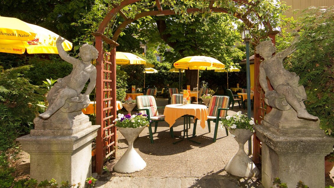 Ein einladender Garten mit einem Tor aus Holz und Steinfiguren. Tische mit gelben Sonnenschirmen sind umgeben von grünen Pflanzen. | © Hendl-Eck - Schmickl