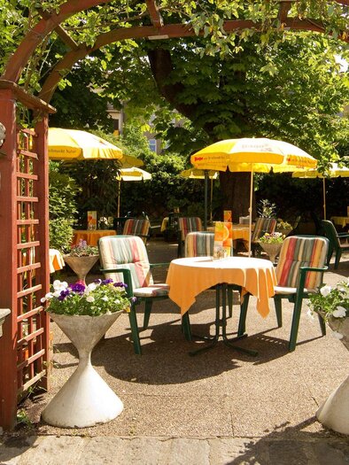 An inviting garden with a gate made of wood and stone figures. Tables with yellow umbrellas are surrounded by green plants. | © Hendl-Eck - Schmickl