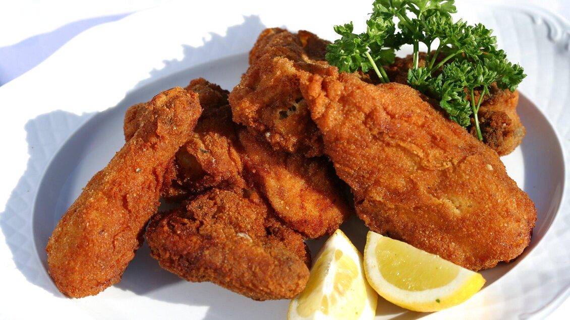 Frittierte Hähnchenschenkel auf einem weißen Teller, garniert mit frischer Petersilie und Zitronenscheiben. Ein appetitliches Gericht, das zum Genießen einlädt. | © Graz Tourismus - Harry Schiffer