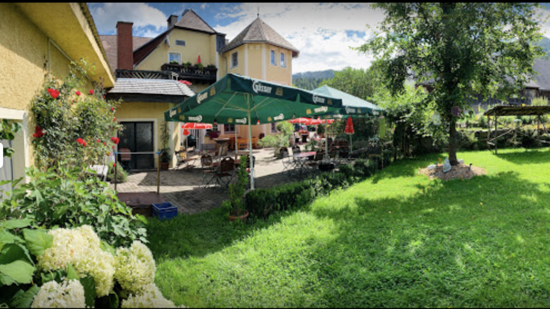 Ein einladender Garten mit grünem Rasen und bunten Blumen. Im Hintergrund steht ein gemütliches Restaurant mit Sonnenschirmen und Tischen. | © Hubinger