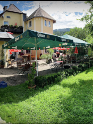 Ein einladender Garten mit grünem Rasen und bunten Blumen. Im Hintergrund steht ein gemütliches Restaurant mit Sonnenschirmen und Tischen. | © Hubinger
