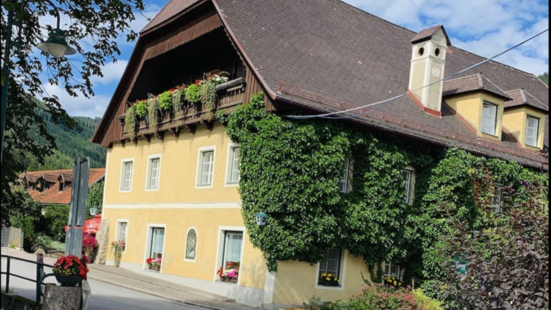 Ein malerisches gelbes Haus, umgeben von üppigem Grün und Blumen. Der Himmel ist blau mit einigen Wolken.