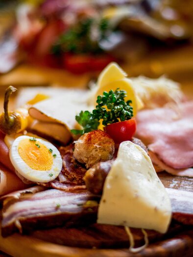 A wooden board with a selection of sausage, cheese, boiled egg, and fresh vegetables. Accompanied by garnishes like parsley and tomatoes.