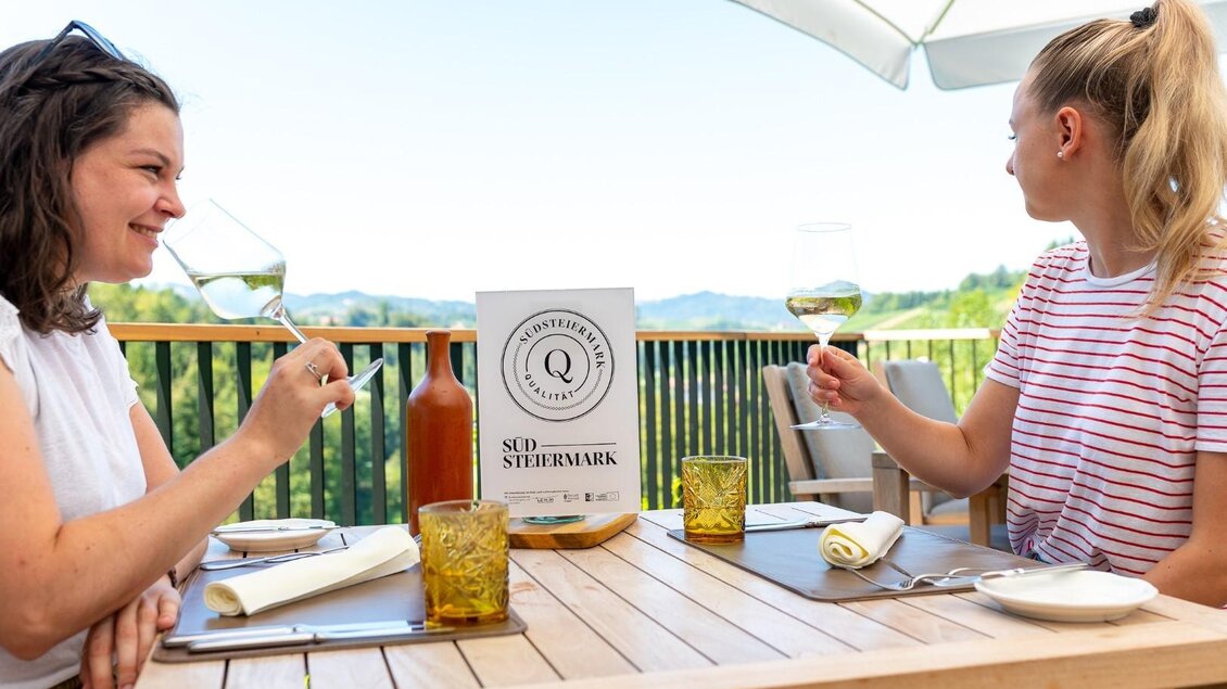 Zwei Frauen sitzen an einem Tisch im Freien und stoßen mit Gläsern an. Im Hintergrund sind grüne Hügel und ein blauer Himmel zu sehen. | © Regionalmanagement Südweststeiermark GmbH/webquartier