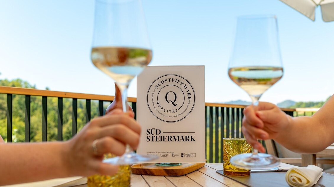 Zwei Personen stoßen mit Weingläsern an. Im Hintergrund ist ein Schild mit der Aufschrift "Südsteiermark" zu sehen. | © Regionalmanagement Südweststeiermark GmbH/webquartier