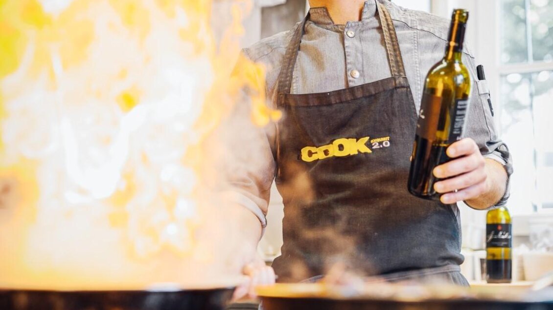 Ein Koch steht in einer Küche und hält eine Flasche Öl in der Hand. Im Hintergrund sieht man Pfannen mit Flammen und Dampf. | © Regionalmanagement Südweststeiermark GmbH/webquartier
