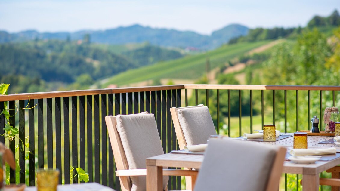 Ein schöner Balkon mit Holzstühlen und einem Tisch. Im Hintergrund erstrecken sich sanfte Hügel und Weinberge unter einem klaren Himmel. | © Regionalmanagement Südweststeiermark GmbH/webquart