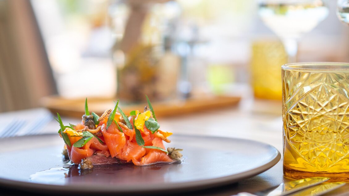 Ein elegantes Gericht mit Lachs, garniert mit frischen Kräutern und Gemüse, auf einem schwarzen Teller. Im Hintergrund ein Getränk in einem goldenen Glas und eine gemütliche Atmosphäre. | © Regionalmanagement Südweststeiermark GmbH/webquart