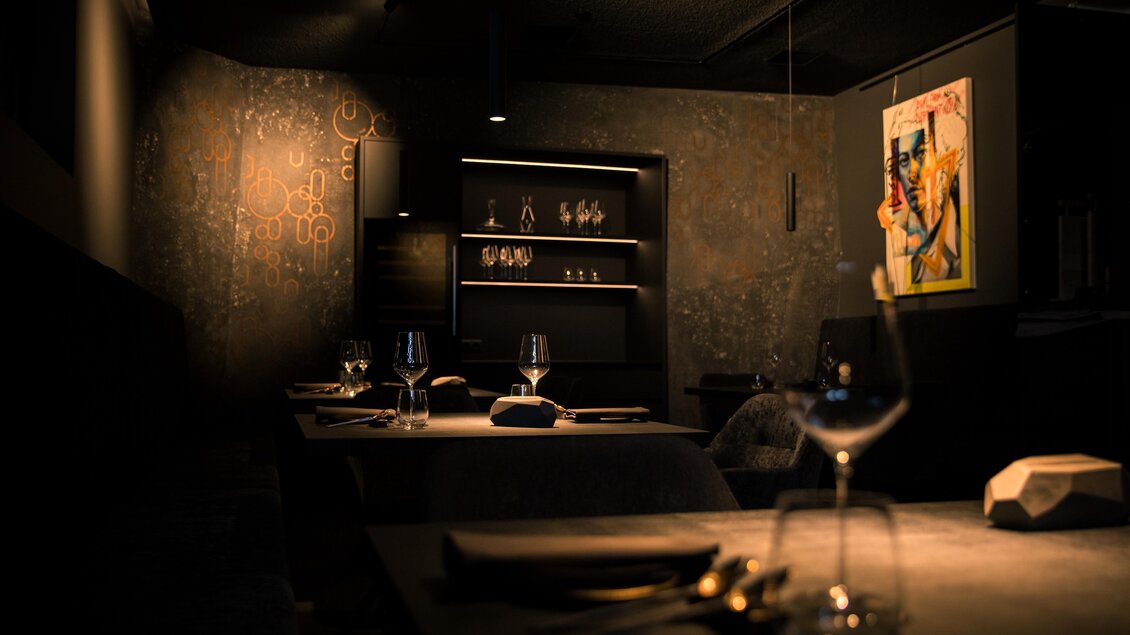 Ein elegantes Restaurant mit gedämpfter Beleuchtung. Auf den Tischen stehen Gläser und Besteck, und eine moderne Kunsttafel ziert die Wand. | © Nadine Geuter - Black Ink Production