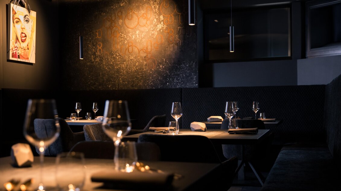 Ein elegantes Restaurant mit dunkler Einrichtung und stilvoll gedeckten Tischen. An der Wand hängt ein Kunstwerk, das das Ambiente ergänzt. | © Nadine Geuter - Black Ink Production