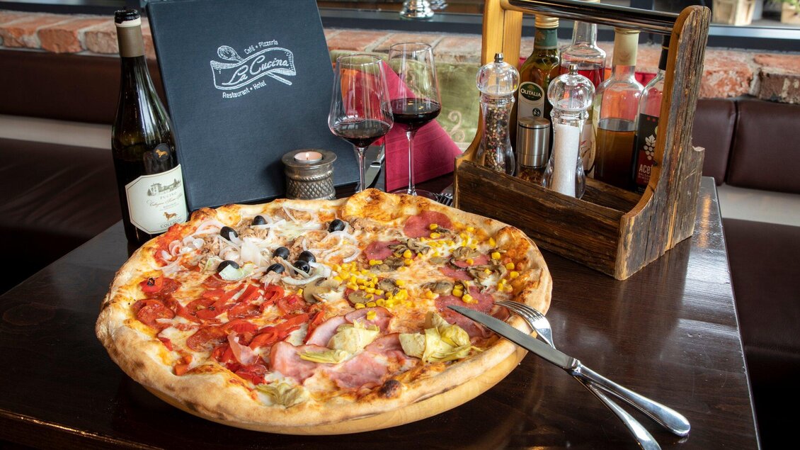 Ein köstlicher Pizza-Teller mit verschiedenen Belägen, darunter Schinken, Oliven und Mais. Im Hintergrund stehen Weinflaschen und Gläser auf einem eleganten Tisch. | © TV Region Graz - Lunghammer