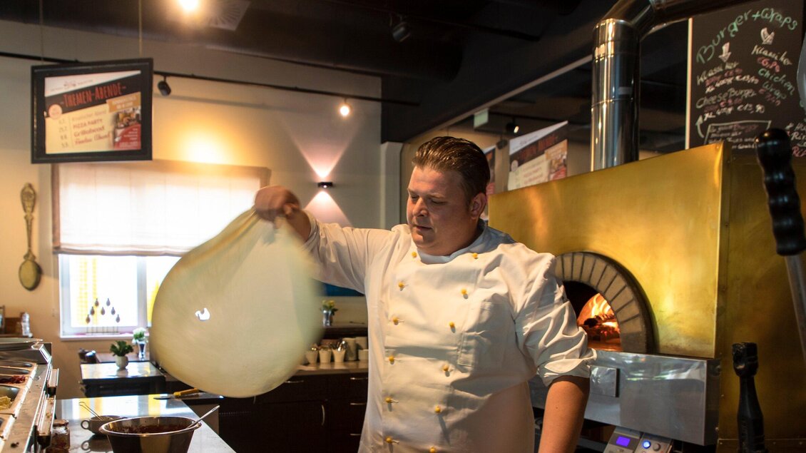 Ein Koch dehnt einen Pizzateig in einer modernen Küche. Im Hintergrund ist ein Pizzaofen und zahlreiche Zutaten sichtbar. | © Region Graz - Lunghammer