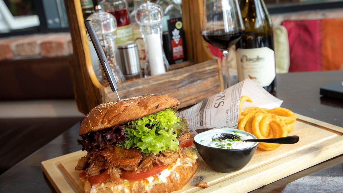 Ein saftiger Burger mit frischem Gemüse, serviert auf einem Holzbrett. Dazu gibt es knusprige Kartoffelringe und eine cremige Sauce. | © TV Region Graz - Lunghammer