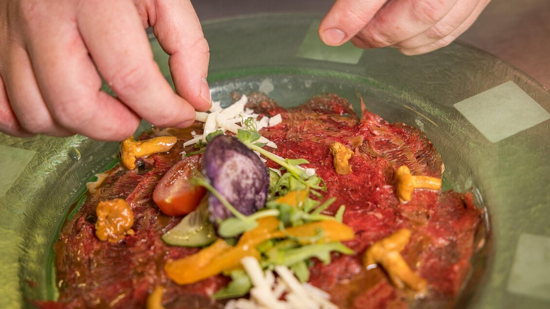 Eine Person dekoriert einen Teller mit rohem Fleisch und frischem Gemüse. Es sind bunte Zutaten wie Pilze, Tomaten und Kräuter sichtbar. | © Netzwerk Kulinarik wildbild
