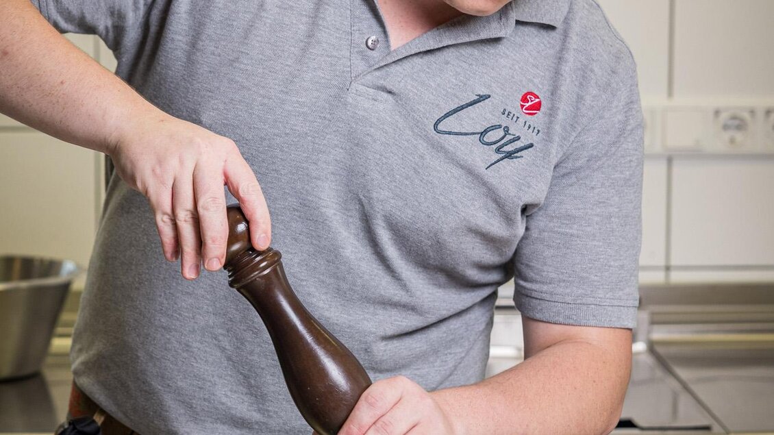 Ein Koch würzt gerade ein Gericht auf einem Teller. Im Hintergrund sind Küchengeräte und eine helle Wand zu sehen. | © Netzwerk Kulinarik wildbild