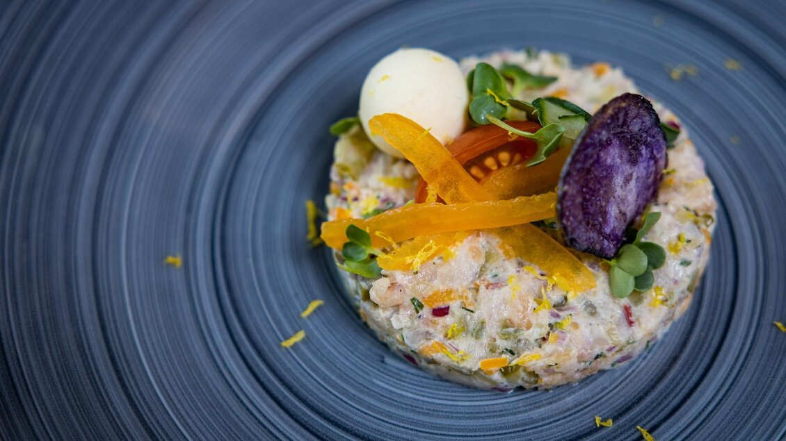Ein kunstvoll angerichteter Teller mit einer delikaten Fischpraline, garniert mit buntem Gemüse und frischen Kräutern. Die lebendigen Farben heben sich auf dem blauen Teller hervor. | © Netzwerk Kulinarik wildbild