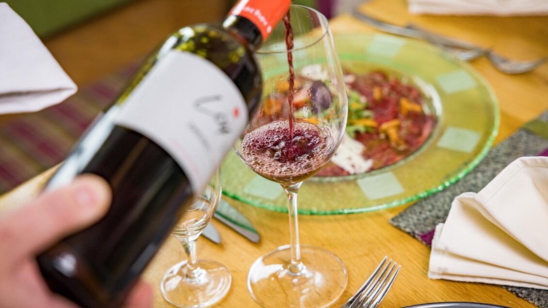 Ein roter Wein wird in ein Glas eingeschenkt, während ein schön angerichteter Teller im Hintergrund sichtbar ist. Die Szene vermittelt ein Gefühl von Genuss und Gemütlichkeit. | © Netzwerk Kulinarik wildbild