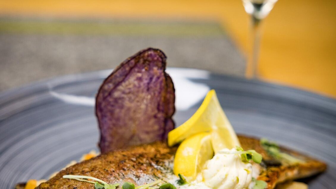 Ein kunstvoll angerichteter Teller mit gebratenem Fisch, buntem Gemüse und einer Zitronenscheibe. Im Hintergrund steht ein Gläser Weißwein. | © Netzwerk Kulinarik wildbild