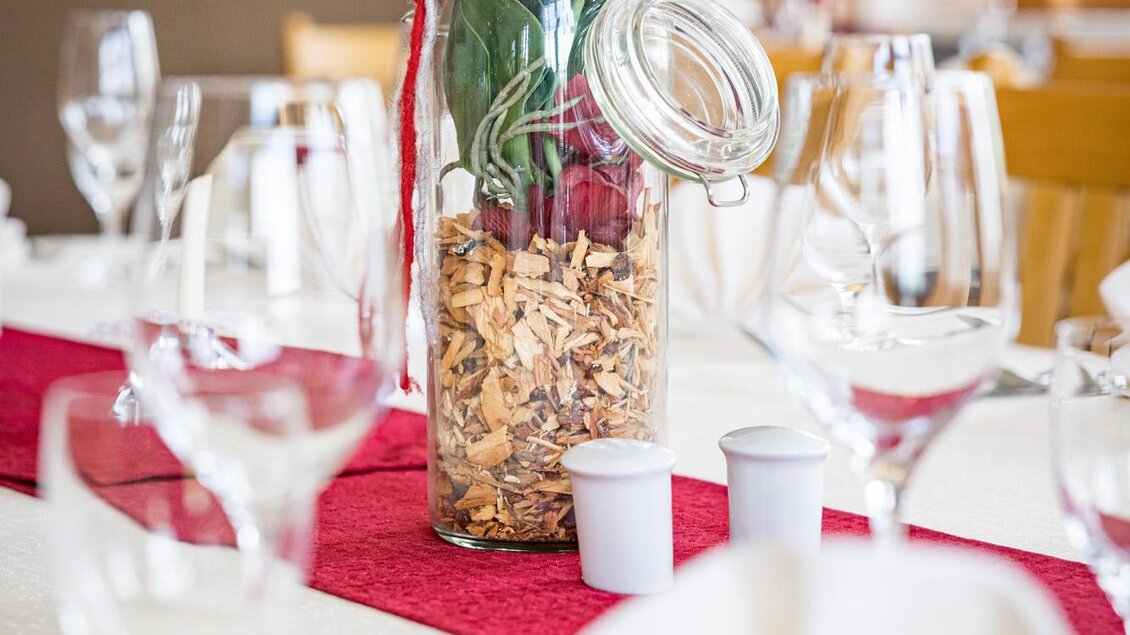 Ein schön dekorierter Tisch mit Gläsern und einem Glas voller Dekoration. Im Glas sind Blumen und Holzstückchen zu sehen, umgeben von einem roten Tischläufer. | © Netzwerk Kulinarik wildbild