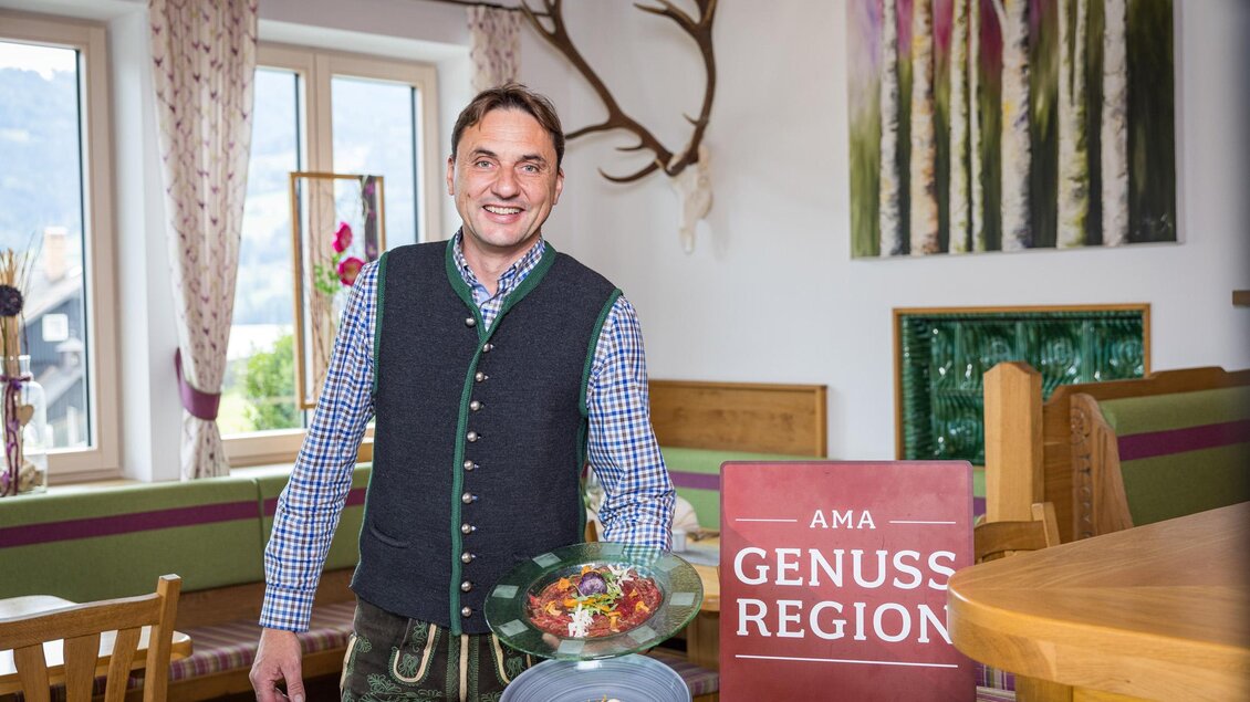 Ein lächelnder Mann in traditioneller Kleidung steht in einem gemütlichen Restaurant. Er hält einen Teller mit regionalem Essen und steht neben einem Schild, das die „Genussregion“ kennzeichnet. | © Netzwerk Kulinarik wildbild