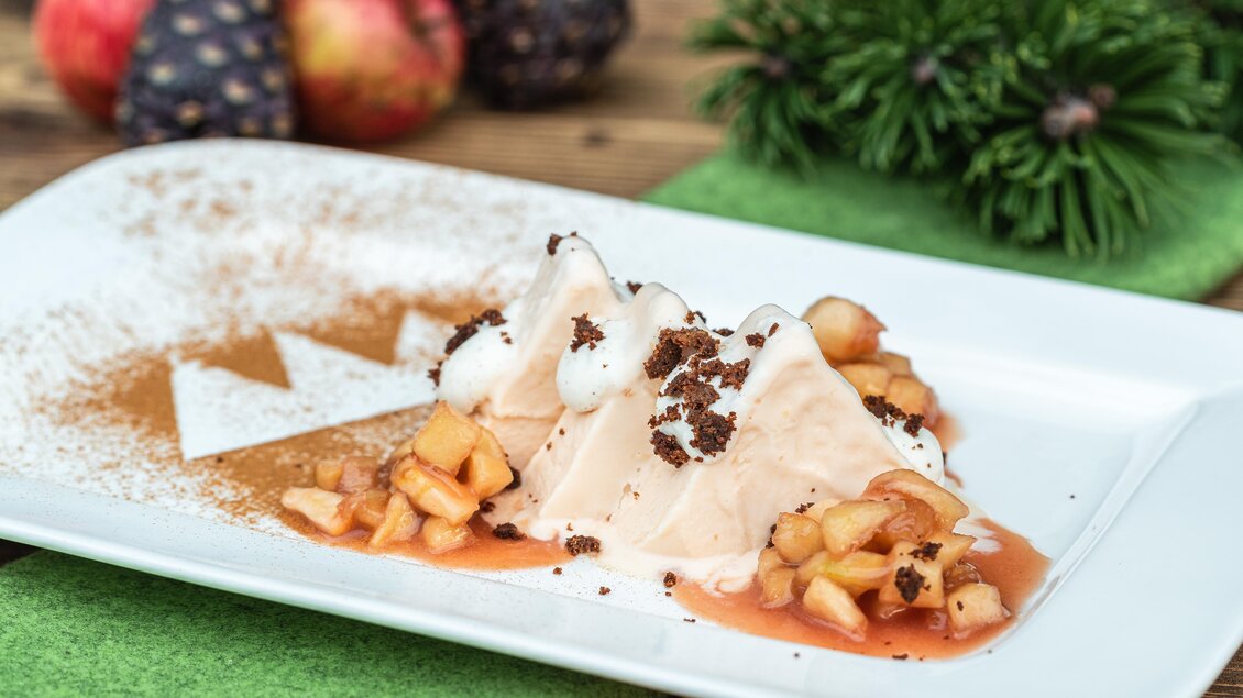 Ein Dessert auf einem weißen Teller mit einer cremigen Schicht, überzogen mit Apfelstücken und Schokoladenkrümeln. Im Hintergrund sind frische Äpfel und Dekorationen zu sehen. | © TVB Schladming-Dachstein/Martin Huber