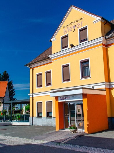 Ein farbenfrohes Restaurant mit gelber Fassade und modernen Fenstern. Die Umgebung ist ruhig mit grünen Bäumen und klar blauem Himmel. | © TV Region Graz - Mias Photoart