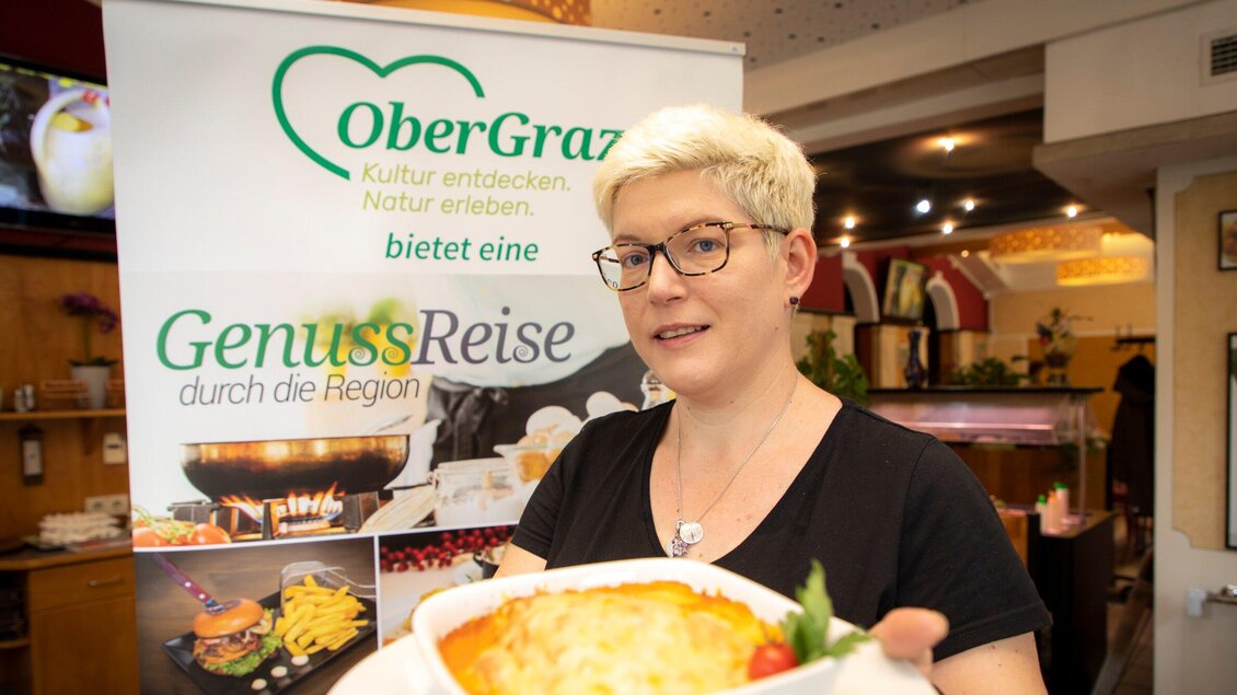 Eine Person hält eine Schüssel mit einem Gericht in der Hand. Im Hintergrund ist ein Banner mit der Aufschrift "OberGraz" und Informationen zur Genussreise zu sehen. | © TV Region Graz - Lunghammer