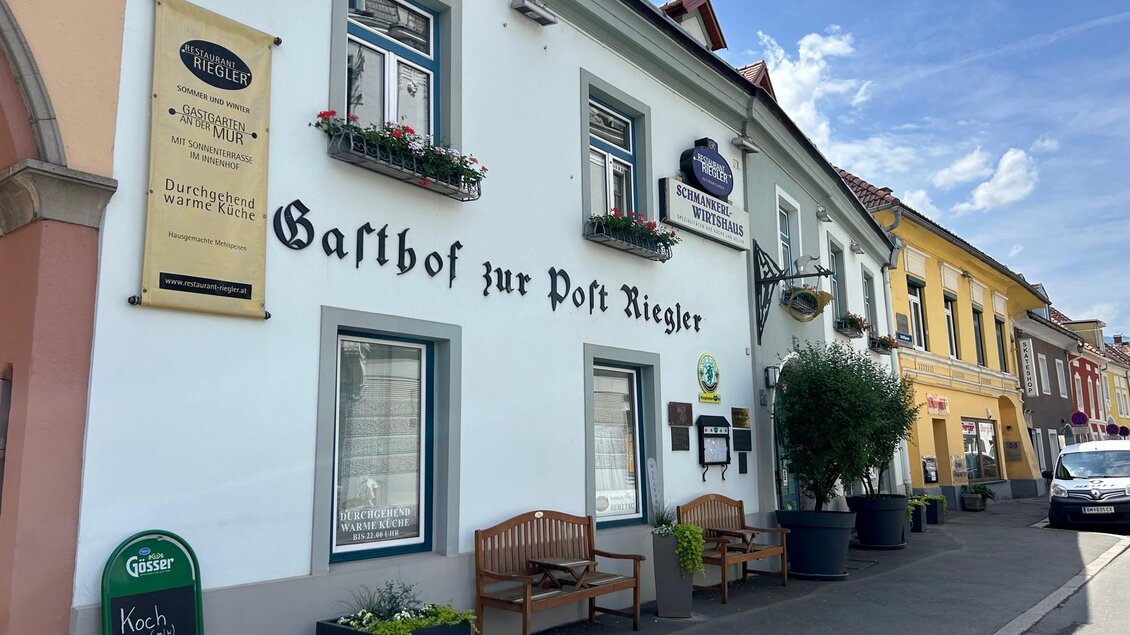 Ein Gasthof mit der Aufschrift "Zur Post Riegler" in einer ruhigen Straße. Die Fassade ist hell, mit Blumenkästen und Bänken im Freien. | © TV Hochsteiermark