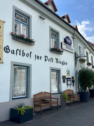A guesthouse with the sign "Zur Post Riegler" on a quiet street. The facade is bright, with flower boxes and benches outside. | © TV Hochsteiermark