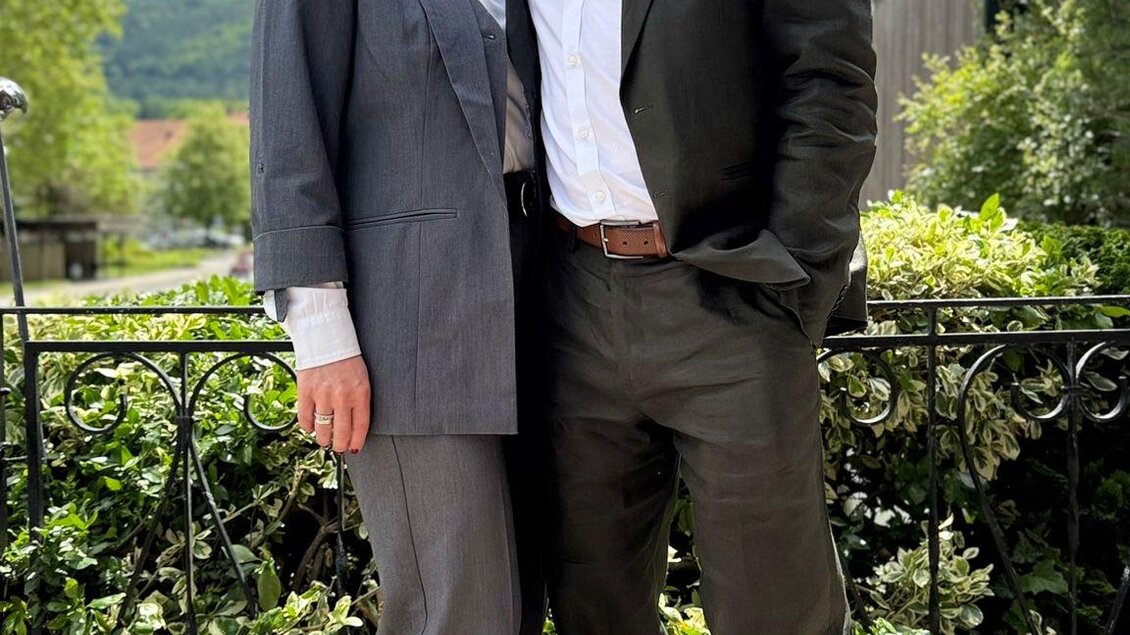 Ein Paar steht lächelnd nebeneinander vor einer grünen Gartenlandschaft. Beide tragen elegante Kleidung und genießen das schöne Wetter. | © Restaurant Riegler