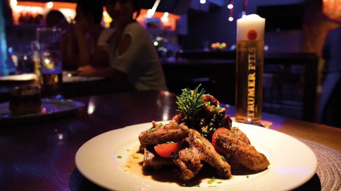 Teller mit gegrillten Hühnerflügeln, Tomaten und Kräutern vor Bierglas in Bar-Atmosphäre. | © Restaurant Rondo