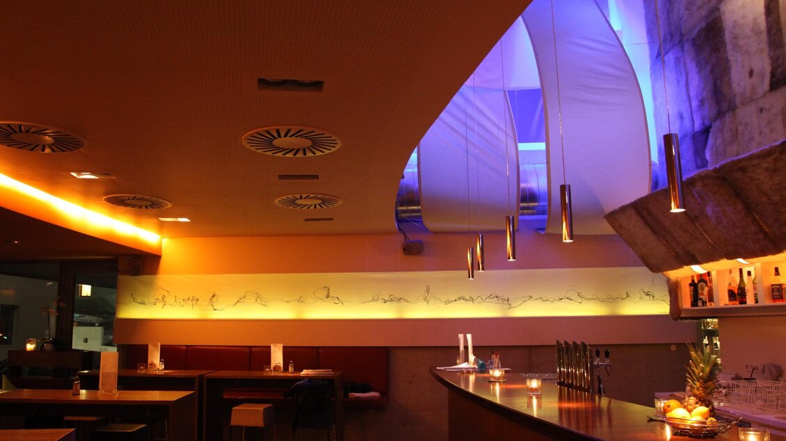 Lange Holzbar mit Kerzen, Obstschale und Hockern vor gelb-blauer Ambientebeleuchtung. | © Restaurant Rondo