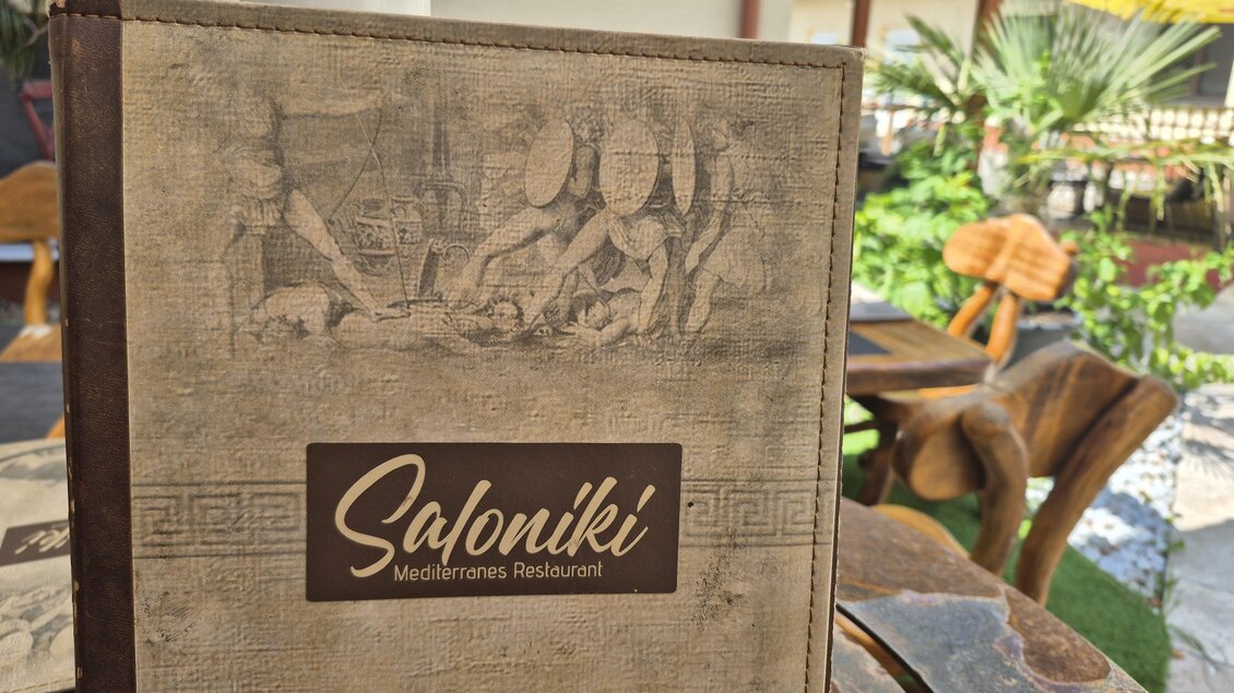 Ein Restaurantmenü mit dem Namen "Saloniki" steht auf einem Tisch. Im Hintergrund ist eine grüne, einladende Terrasse zu sehen. | © TV Hochsteiermark