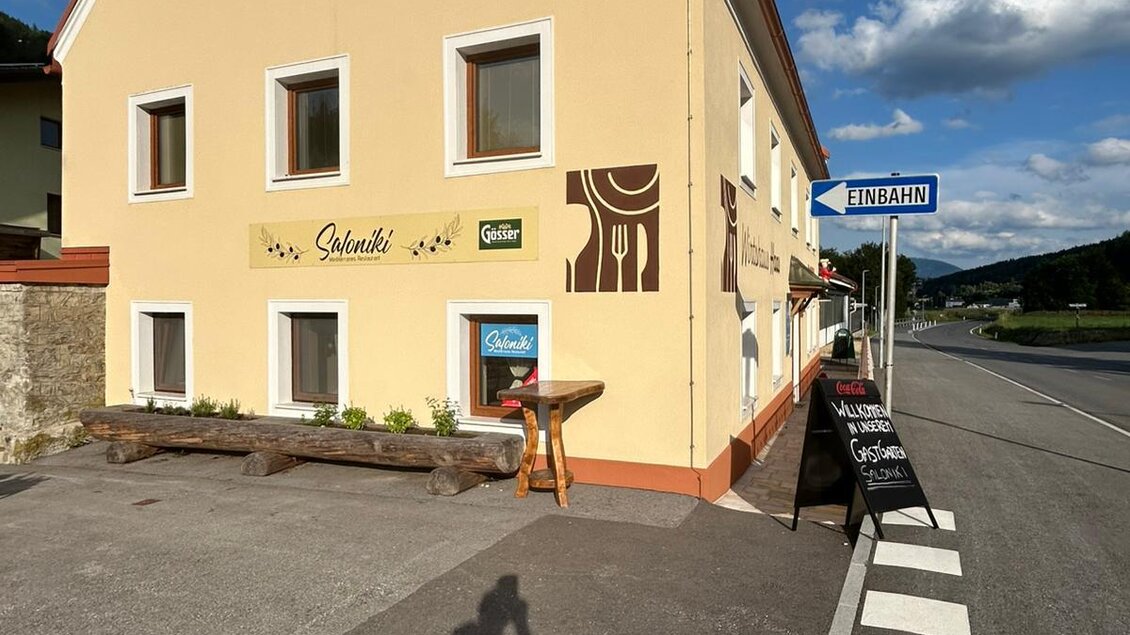 Ein farbenfrohes Gebäude an einer Straße mit einem Schild und einem Tisch im Vordergrund. Am Straßenrand stehen Hinweisschilder und es ist ein klarer Himmel zu sehen. | © TV Hochsteiermark