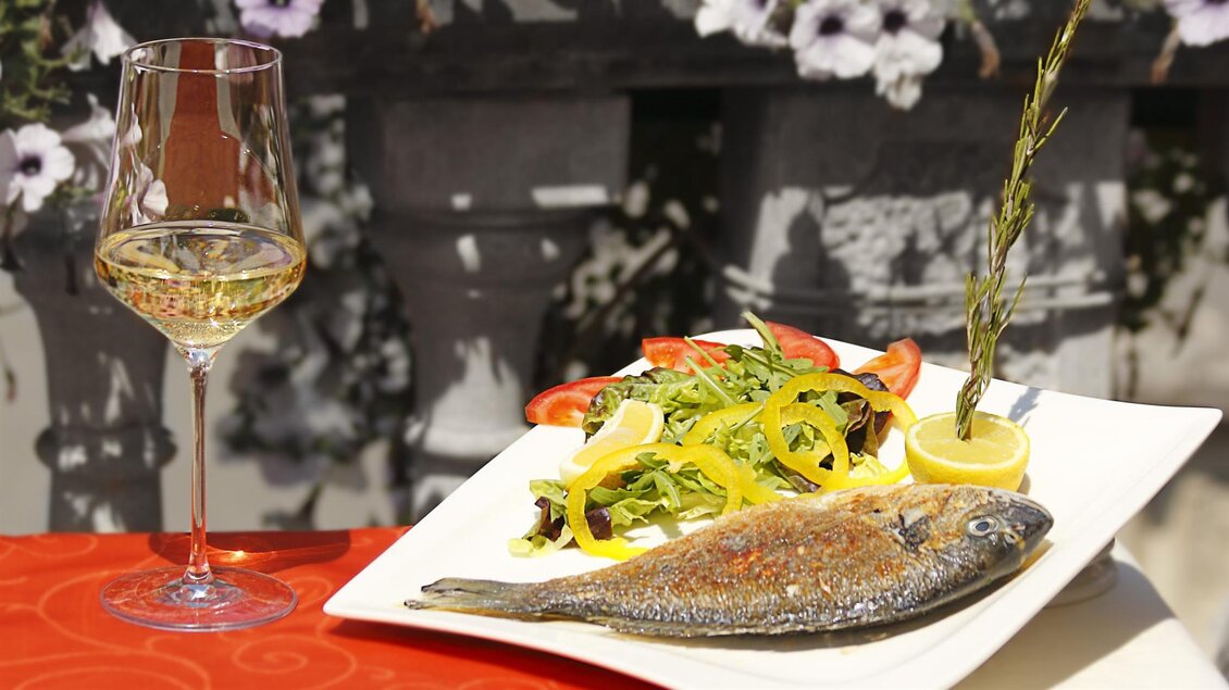 Ein lecker gegrillter Fisch auf einem Teller, begleitet von frischem Salat und einer Zitrone. Ein Glas Weißwein steht daneben. | © San Marco Hotel GmbH