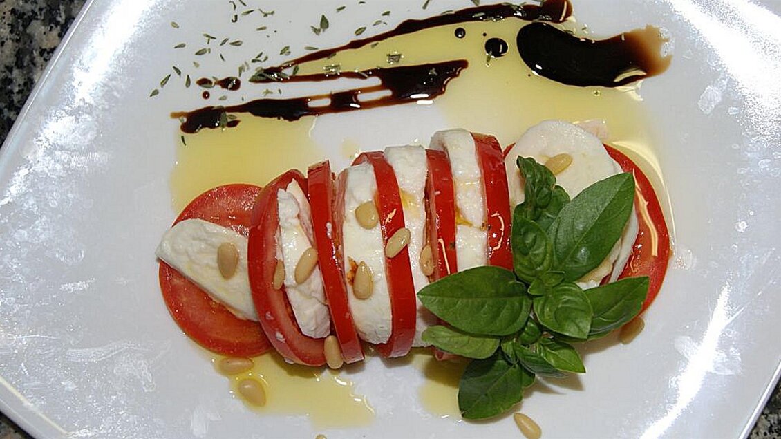 Ein Teller mit frischer Caprese, bestehend aus Tomaten, Mozzarella, Basilikum und Pinienkernen. Darüber ist Balsamico und Öl dekoriert. | © San Marco Hotel GmbH