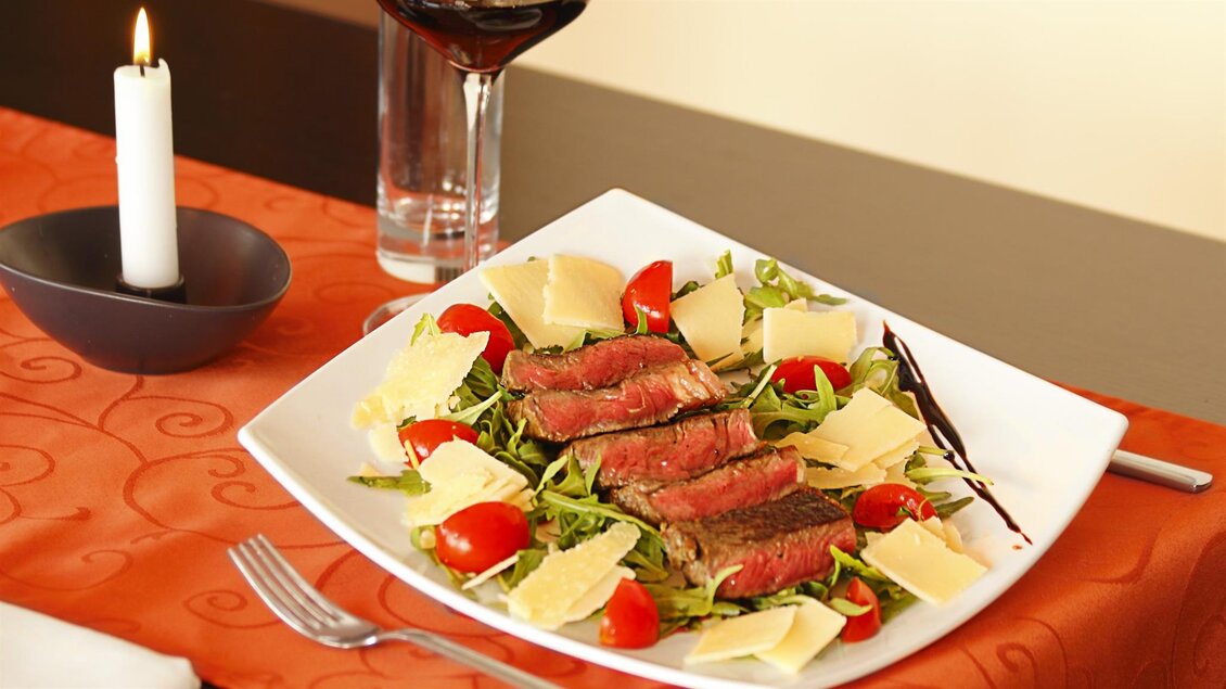 Ein Teller mit frischem Salat, saftigem Steak und Käsestücken. Im Hintergrund sind ein Glas Rotwein und eine Kerze zu sehen. | © San Marco Hotel GmbH