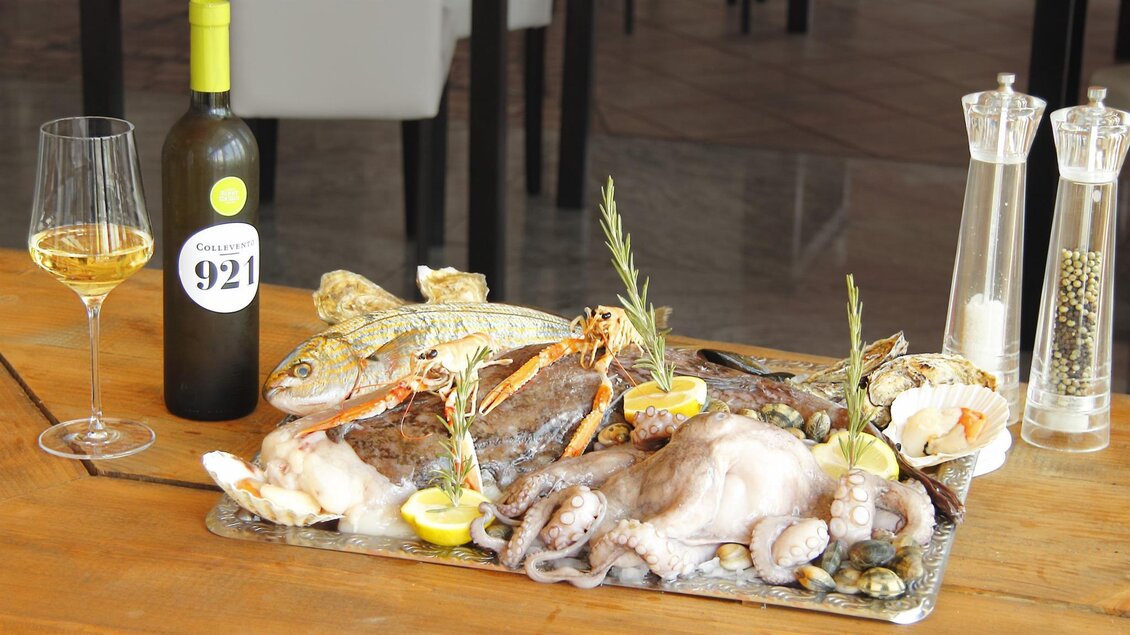 Ein köstliches Meeresfrüchte-Gericht auf einem Blech, garniert mit Zitronen und Kräutern. Dazu ein Glas Weißwein und eine Flasche Wein auf dem Tisch. | © San Marco Hotel GmbH