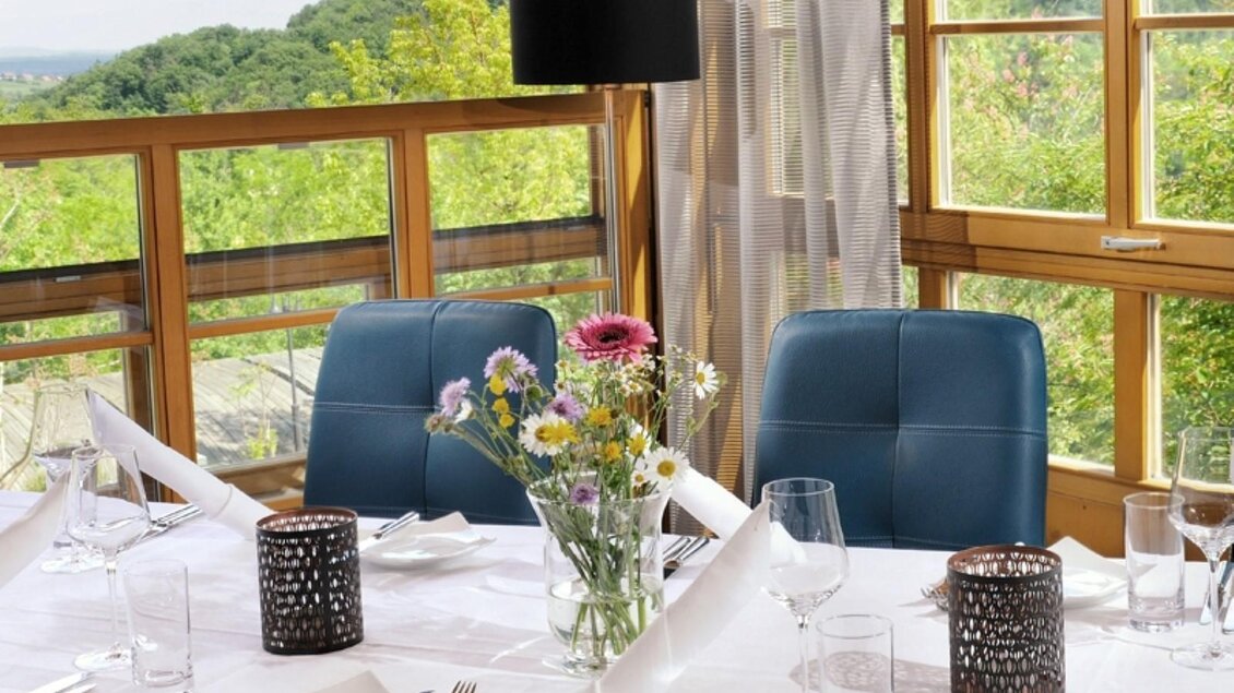 Ein eleganter Tisch in einem Restaurant mit Blick auf die Natur. Blumen und Kerzen sorgen für eine einladende Atmosphäre. | © Sattler