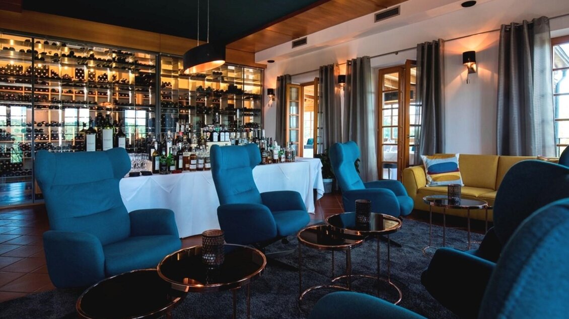 Ein stilvolles Wohnzimmer mit blauen Sesseln und eleganten Tischchen. Im Hintergrund ist eine gut sortierte Bar zu sehen. | © Sattler