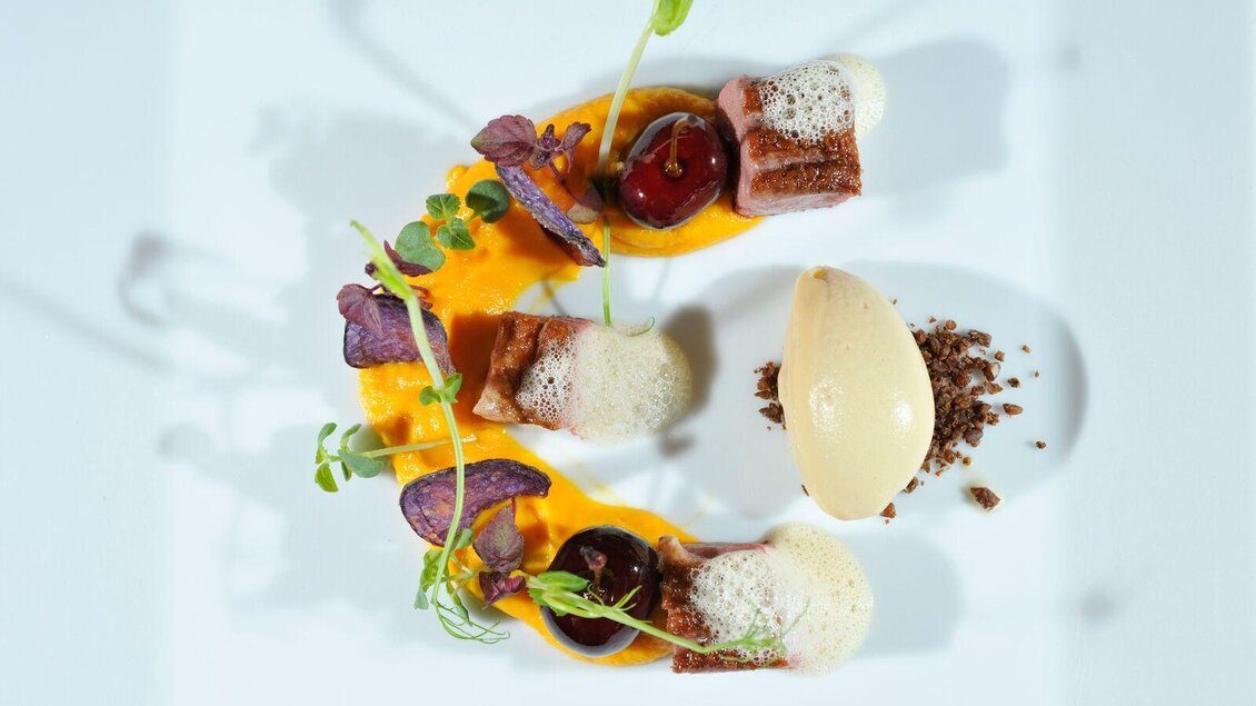 Ein kunstvoll angerichtetes Gericht mit Fleischstücken, buntem Gemüse und einer cremigen Sauce. Dazu gibt es eine Portion Eiscreme und essbare Kräuter zur Dekoration. | © Sattler