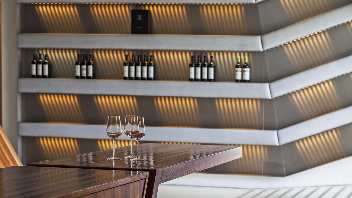 Ein eleganter Innenraum mit einem modernen Holzstahltisch und einer stilvollen Wand mit beleuchteten Weinflaschen. Der Ort strahlt eine angenehme Atmosphäre aus. | © Sattler
