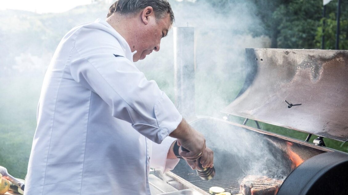 Ein Koch grillt Fleisch und Gemüse im Freien. Rauch steigt auf und das Wetter sieht schön aus. | © Sattler